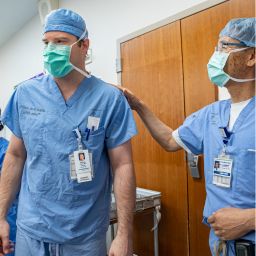 Two men in blue surgery scrubs, masks and bonnets. Man on right has his right hand on other man's left shoulder.