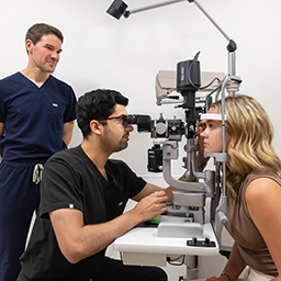 Young man examining young female patient using the slit lamp while a man observes in the background.