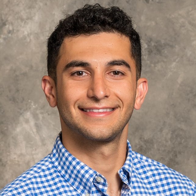 Smiling young man with short dark curly hair, wearing a blue and white plaid shirt.