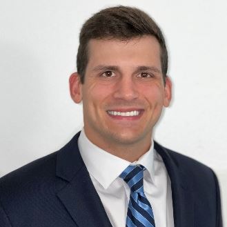 Smiling young man with short brown hair, wearing a dark suit, white dress shirt, and blue striped tie.