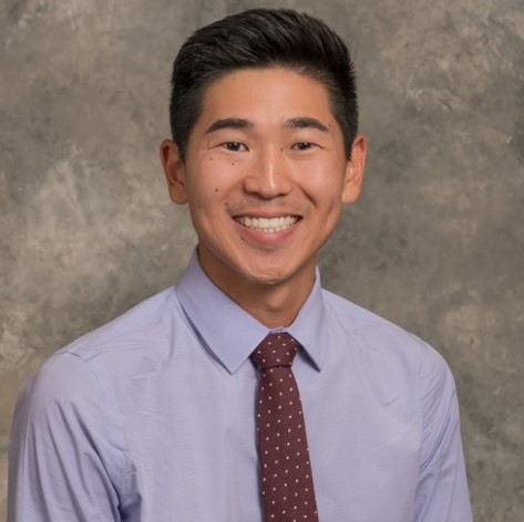 Smiling young man with short dark hair, wearing a purple dress shirt with a burgandy tie.