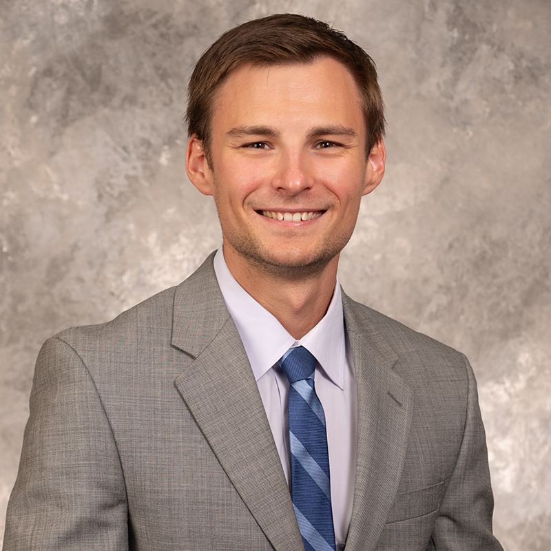 Smiling young man with short brown hair, wearing a brown suit and blue dress shirt.