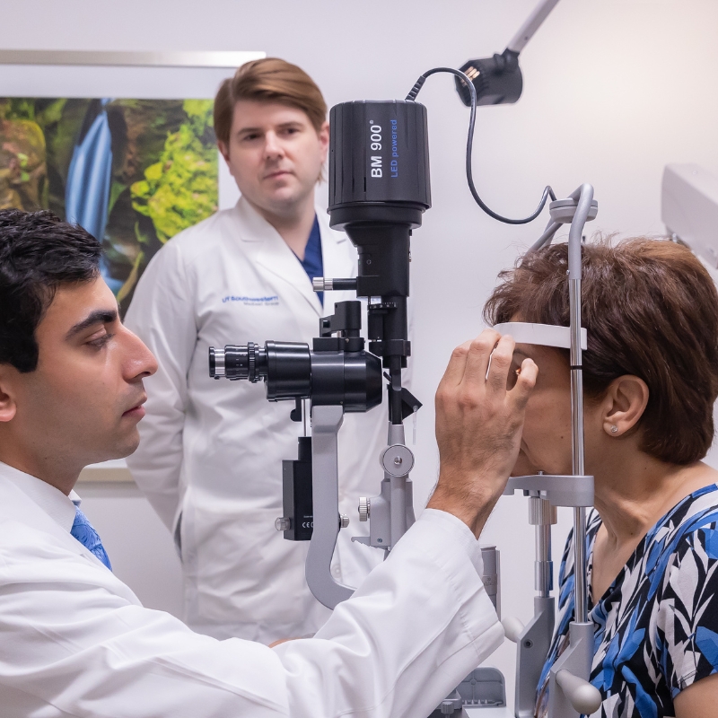 Doctor with black hair and wearing white lab coat examining eye of female patient while another doctor with light brown hair and wearing a white lab coat looks on.