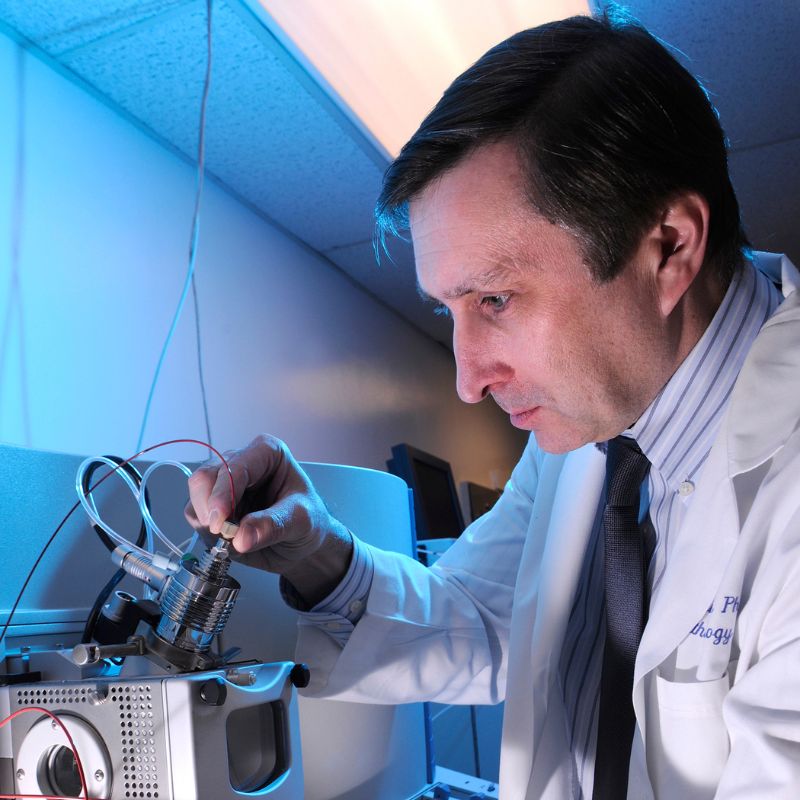 Man with brown hair wearing white lab coat preparing to peer into a piece of ophthalmic lab equipment