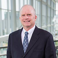 Slightly smiling with receding gray hair, wearing a dark suit, white shirt, and blue patterned tie.