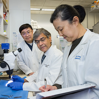 Two men with dark hair and glasses wearing white lab coats looking at what a woman with dark hair wearing a white lab coat is writing in her notebook.