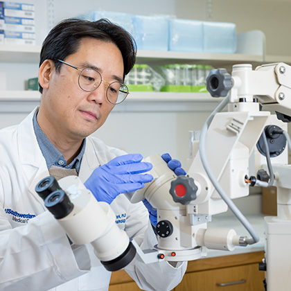 Man with black hair and glasses wearing a white lab coat is examining a lens on a piece of large ophthalmic lab equipment.