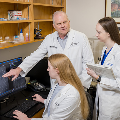 Man with gray hair wearing a white lab coat pointing at data on a computer screen while speaking with two young women in white lab coats.