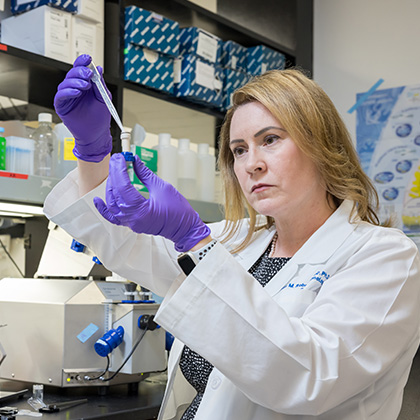 Woman with dark blonde hair wearing purple gloves and a white lab coat transferring liquid into a small vial with a dropper.