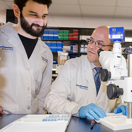 Young man with black hair and beard wearing a white lab coat standing to the right of a man in a white lab coat with a bald head and glasses seated at a microscope and looking to his right.