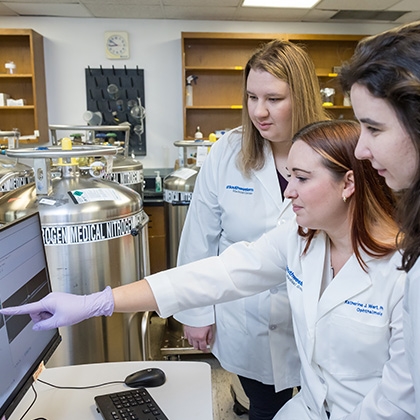 Three young women in white lab coats looking at a computer screen as the woman in the middle points at data.