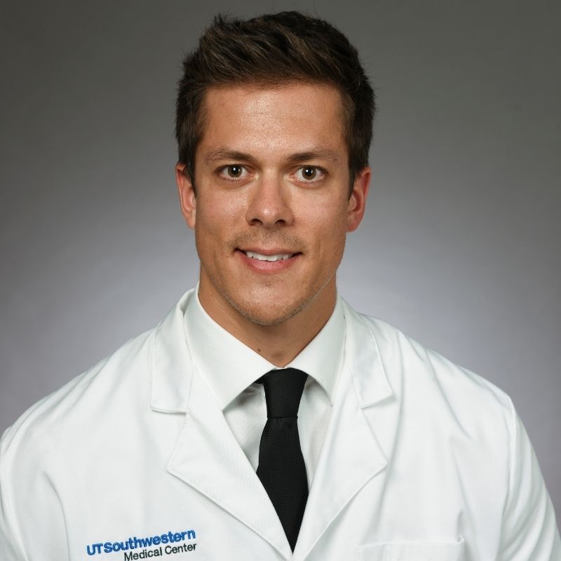 Smiling man with brown hair wearing a white dress shirt, dark blue tie, and lab coat.