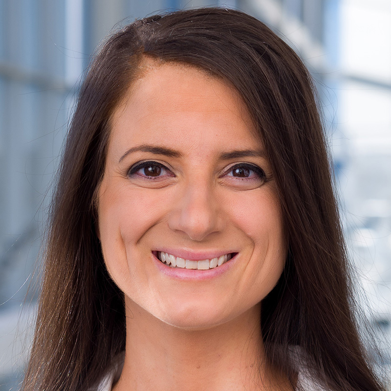 Smiling woman with long brown hair wearing black blouse and white lab coat.