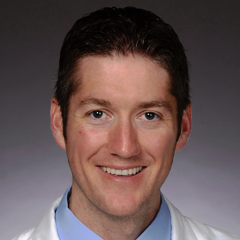 Smiling man with brown hair wearing blue dress shirt, striped tie and white lab coat