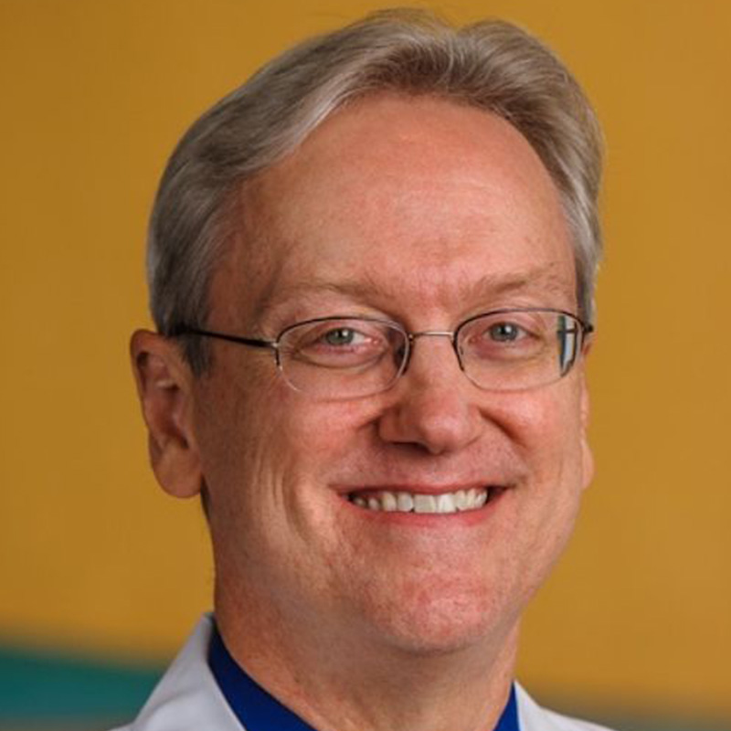 Smiling man with rimless glasses and blonde-grey hair wearing a dark blue dress shirt and tie and white lab coat against a colorful yellow background.