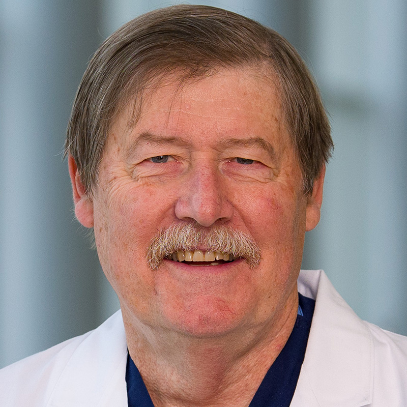 Smiling man with grey hair and mustache wearing dark blue scrubs and a white lab coat.