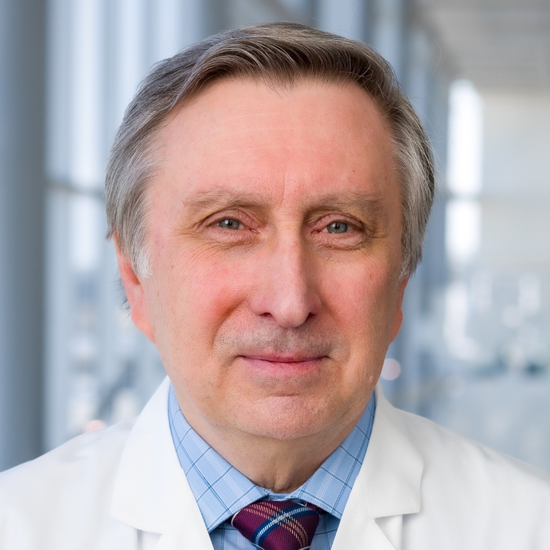 Smiling man with gray hair weating a blue dress shirt, red-striped tie and white lab coat