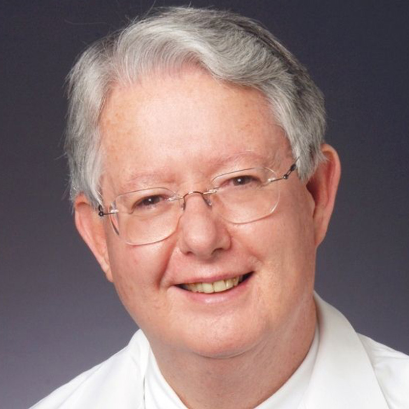 Smiling man with rimless glasses and white hair wearing a white dress shirt, colorful patterned tie, and a white lab coat.