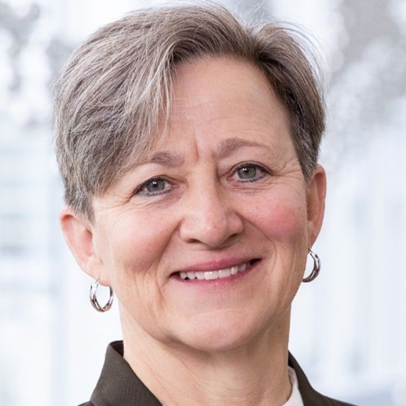 Smiling woman with short gray hair wearing small silver hoop earrings, a white blouse, and brown blaze