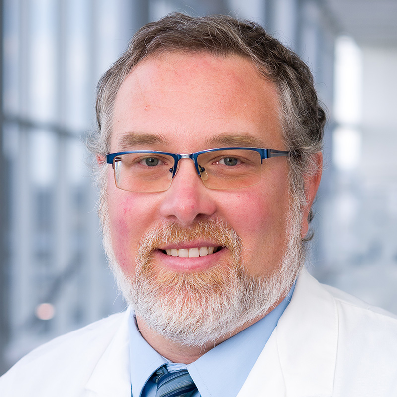 Smiling man with bray hair, white beard and mustache wearing blue-rimmed glasses, a light blue shirt, blue-striped tie, and white lab coat