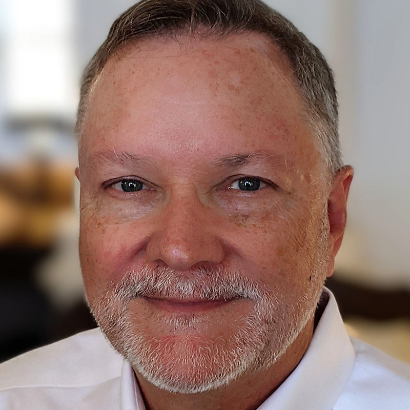 Slightly smiling man with brown gray hair and a gray and white goatee wearing a white dress shirt