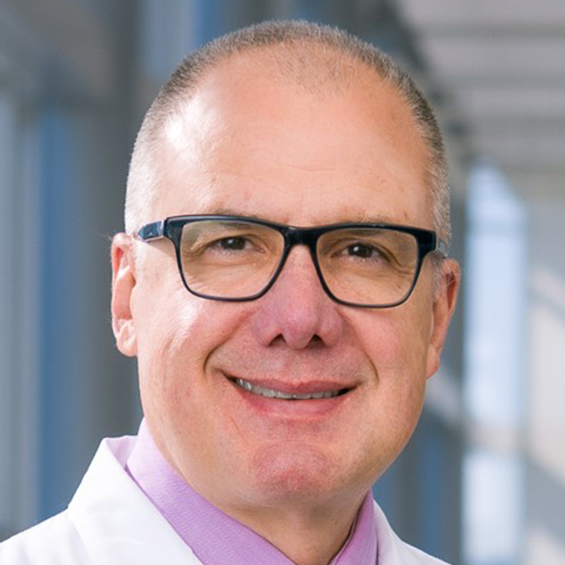 Smiling man with gray hair, wearing a white lab coat and dark-rimmed glasses.