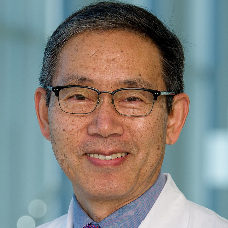 Smiling man with black and gray hair wearing black-rimmed glasses, a light blue dress shirt, purplle tie, and white lab coat