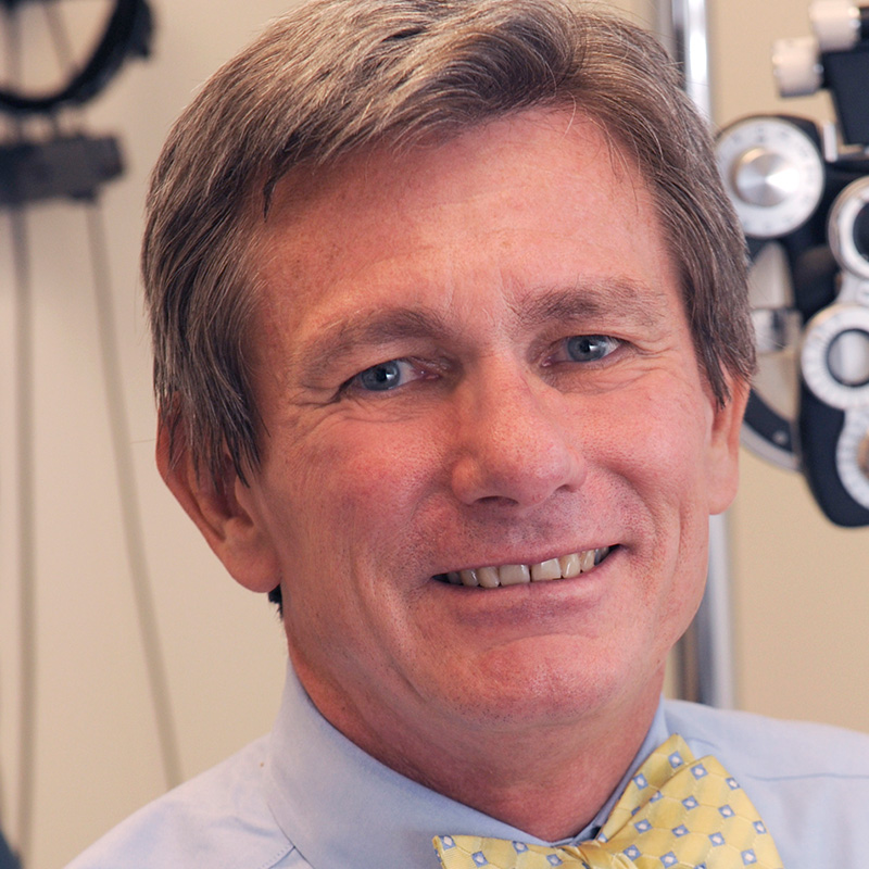 Smiling man with brown-gray hair in front of a slit lamp wearing a light blue tie and yellow and blue bow tie.