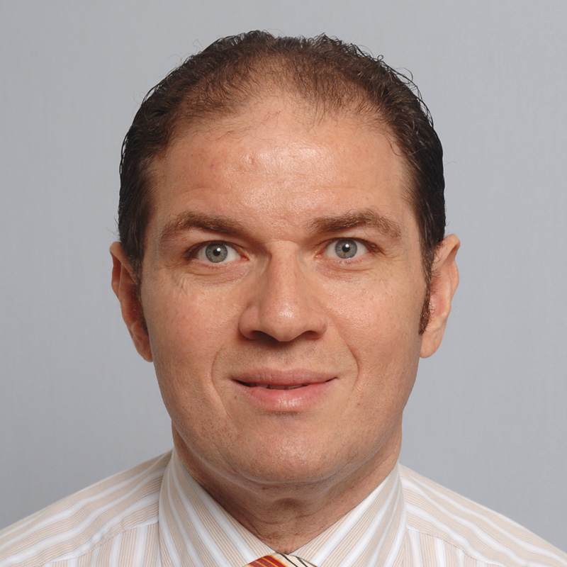 Slightly smiling man with black hair wearing a beige and white-striped dress shirt and striped tie.