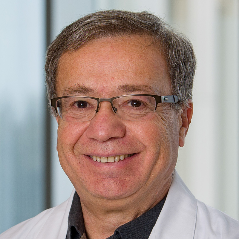 Smiling man with gray hair wearing brown-rimmed glasses, a blue polo shirt and white lab coat.