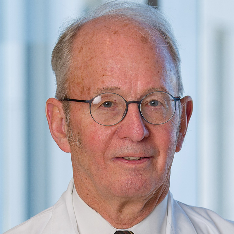 Smiling man with white hair waring round metal framed glasses, white shirt, brown paisley tie and white lab coat.