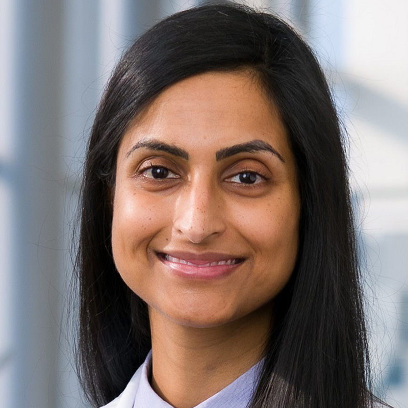 Smiling woman with long black hair wearing a light blue blouse and white lab coat.