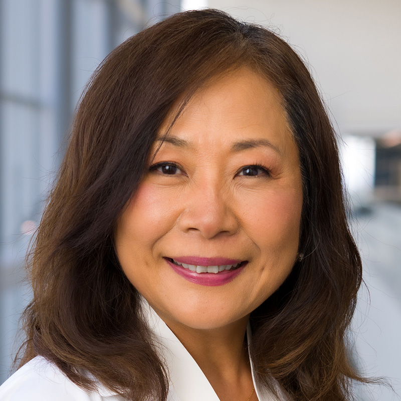 Smiling woman with black-brown hair wearing a white blouse and white lab coat.