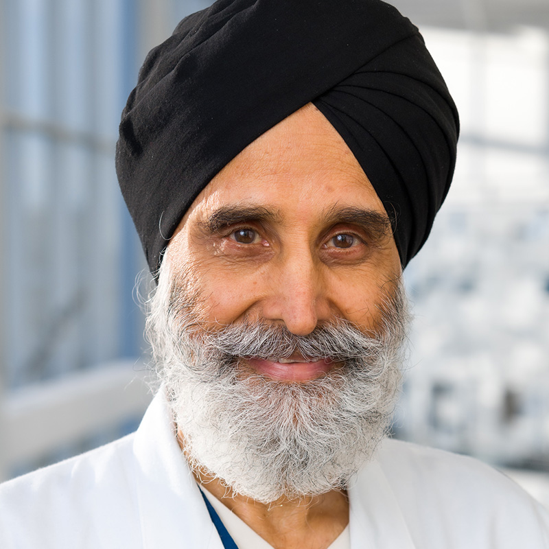 Smiling man with gray mustache and beard wearing a black turban, blue scrubs and a white lab coat.