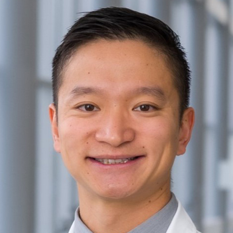 Smiling man with black hair wearing a gray dress shirt, blue and gray striped tie, and white lab coat.