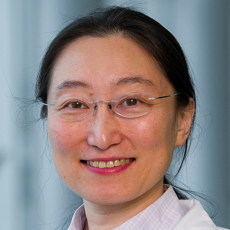 Smiling woman with black hair tied back in a pony tail wearing frameless glasses, a pink-checked blouse and white lab coat.