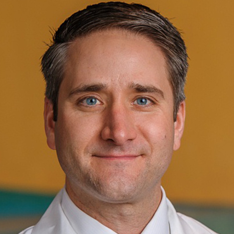 Smiling man with brown hair wearing a white dress shirt, yellow patterned tie, and white lab coat.