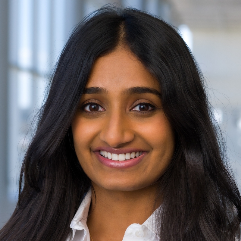 Smiling young woman with long, wavy dark brown hair wearing a white lab coat.