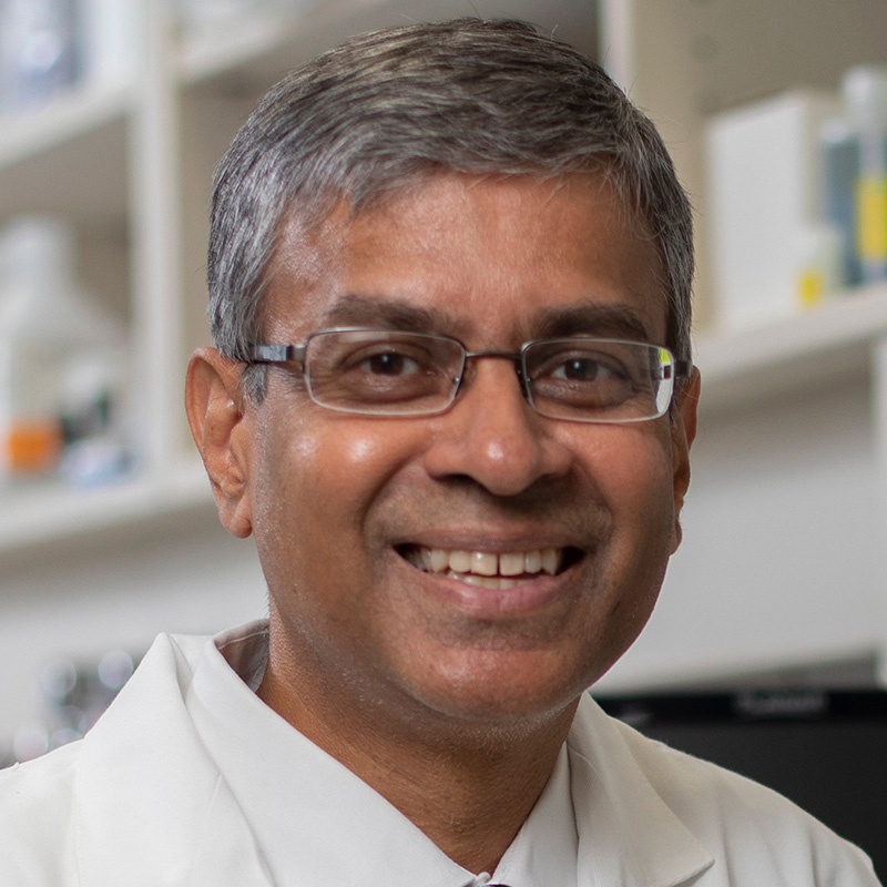 Smiling man with black and gray hair wearing silver framed glasses, a white dress shirt, blue striped tie, and white lab coat