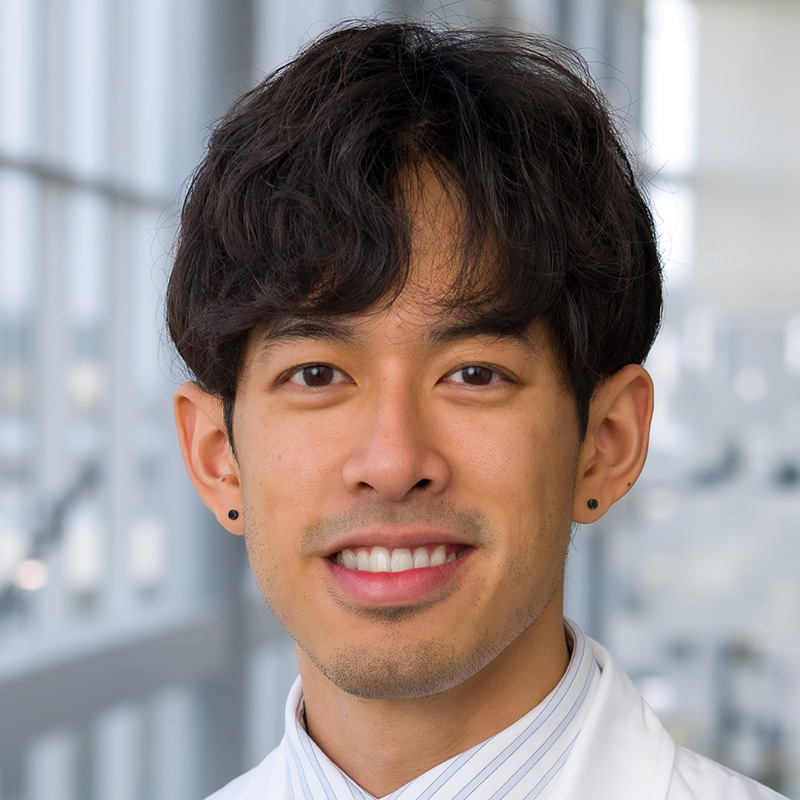 Smiling man with black hair wearing black button earrings, a white and gray striped dress shirt, dark blue tie, and white lab coat.