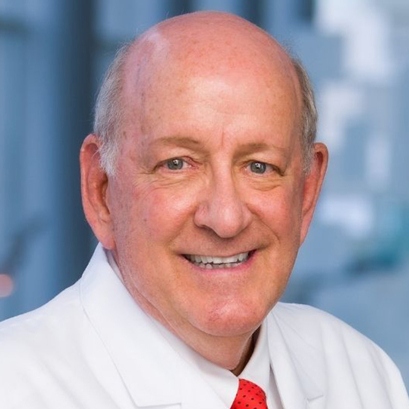 Smiling, mostly bald man with white-gray hair on the side wearing a white dress shirt, red tie, and white lab coat.