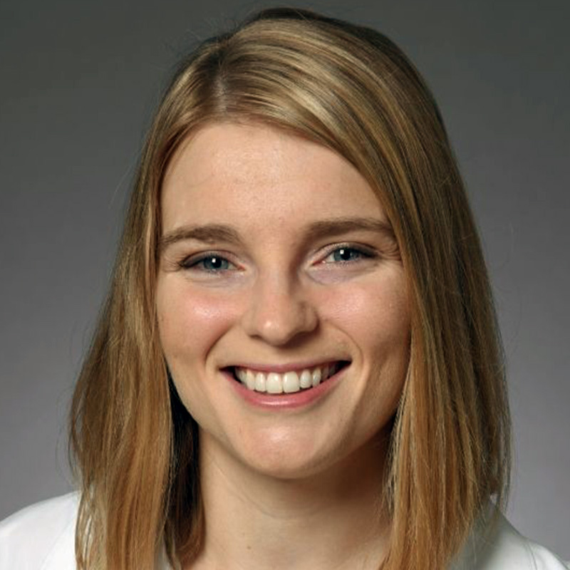 Smiling woman with shoulder-length blonde hair wearing a burgundy blouse and white lab coat.