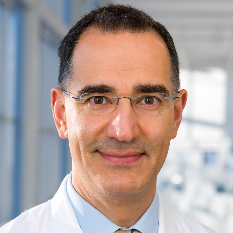 Smiling man with black hair wearing rimless glasses, a light blue dress shirt, striped tie, and white lab coat.