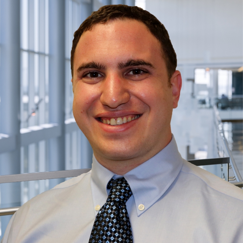 Smiling young man with brown curly hair and brown eyes wearing a light gray dress shirt and dark blue patterned tie.