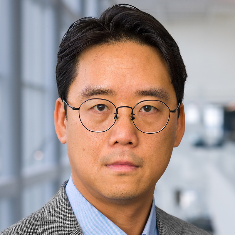 Unsmiling man with black hair and round glasses wearing a light blue dress shirt and black and white tweed sports coat.