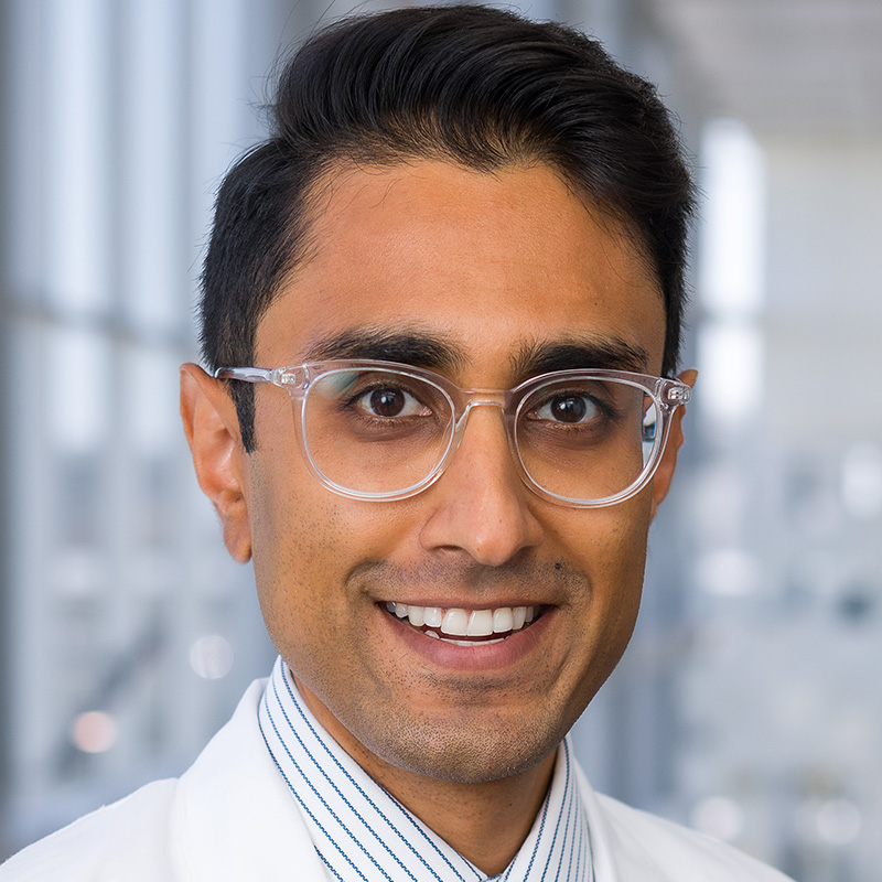 Smiling man with black hair wearing clear acrylic round glasses, a blue and white striped dress shirt, dark blue patterned tie, and white lab coat.