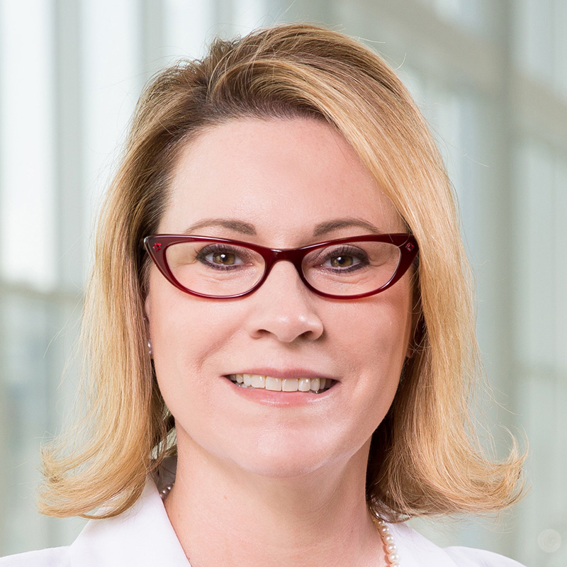 Smiling woman with blonde hair wearing red cat-eye glasses, a pearl necklace, a blue and white striped bouse, and a white lab coat.