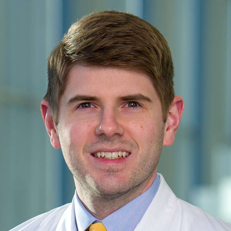 Smiling man with brown hair wearing a light blue dress shirt, yellow tie, and white lab coat.