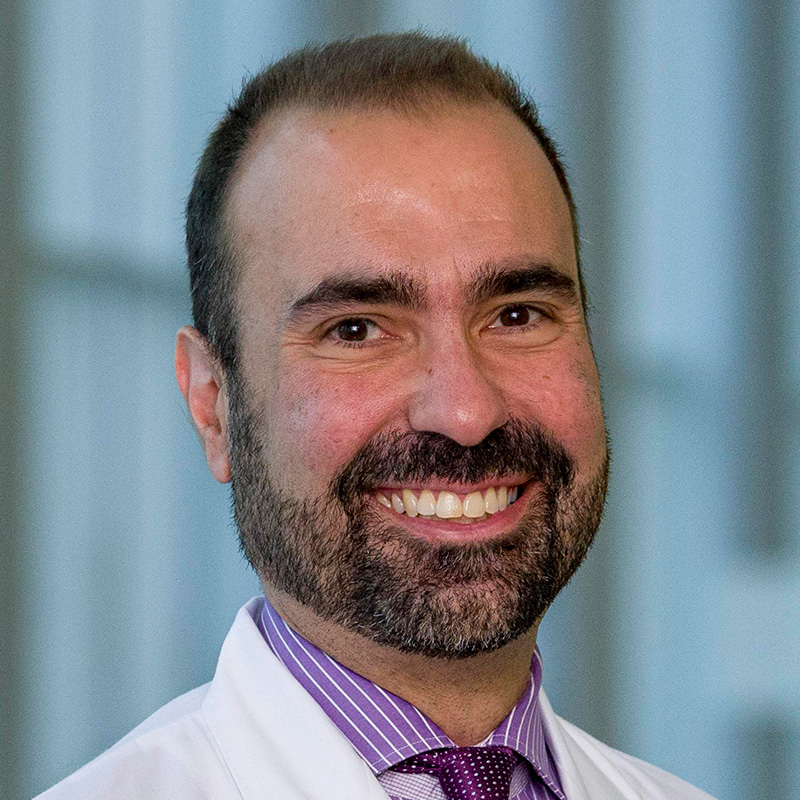 Smiling man with black hair, goatee, and mustache wearing a purple dress shirt and tie, and white lab coat.