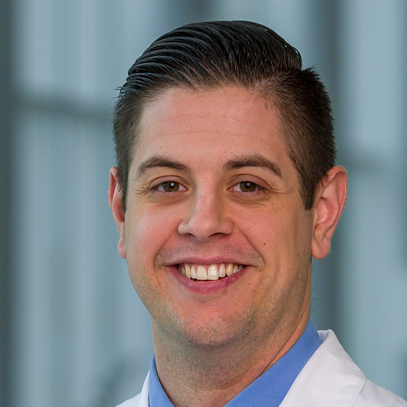 Smiling man with brown hair wearing a blue dress shirt, blue striped tie and white lab coat.